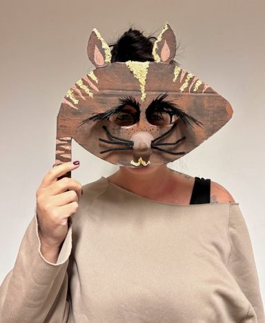 Frau hält eine selbstgemachte Tiermaske mit Katzenmotiv vor ihr Gesicht.
