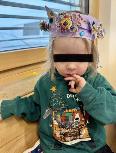 Kleines Kind mit buntem Hut und grünem Pullover, sitzt nachdenklich und hält den Finger am Mund.