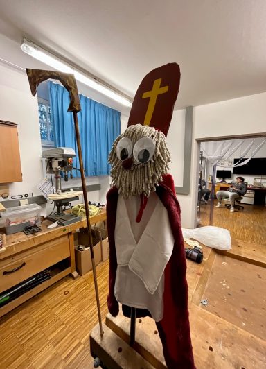 Figur eines heiligen Mannes mit rotem Umhang und Stab, auf einem Sockel stehend.