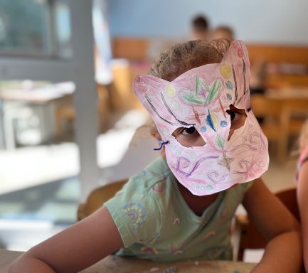 Kind trägt eine bunte, selbstgemachte Maske und sitzt an einem Tisch.